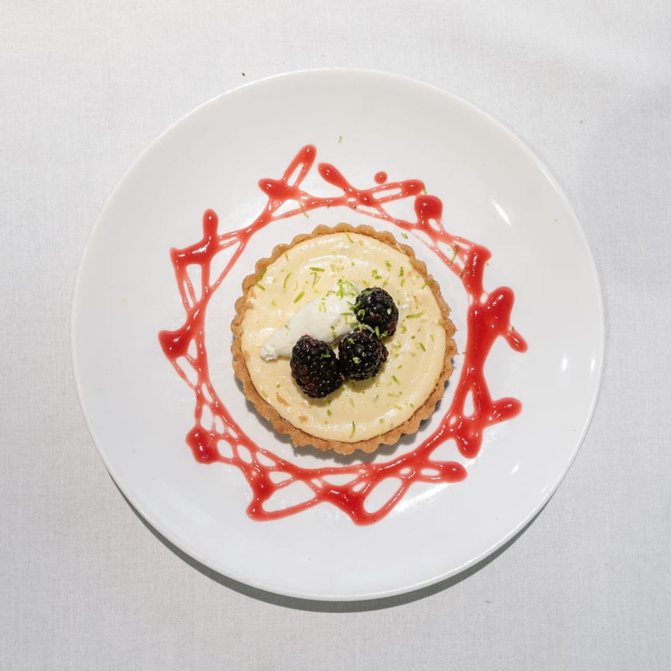 Key lime tart with whipped cream, blackberries, lime zest, and raspberry drizzle on a white plate.
