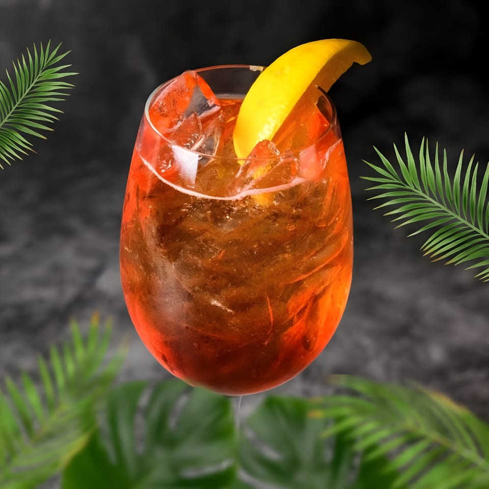 A vibrant orange cocktail with ice and a lemon wedge, surrounded by green palm leaves.