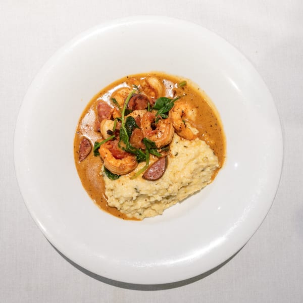 Shrimp and grits with sausage and spinach in a rich sauce on a white plate.