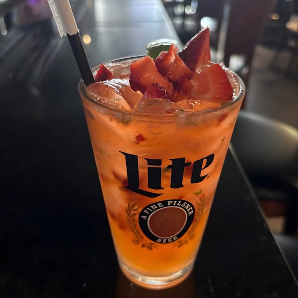 A tall glass of orange-colored drink with ice, strawberries, and a straw on a dark bar.