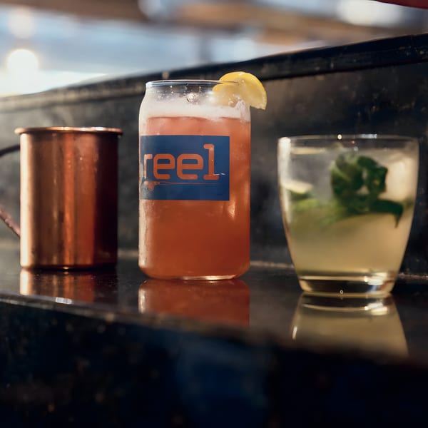 Three drinks on a dark bar: a copper mug, a Reel-branded pink cocktail, and a mojito.